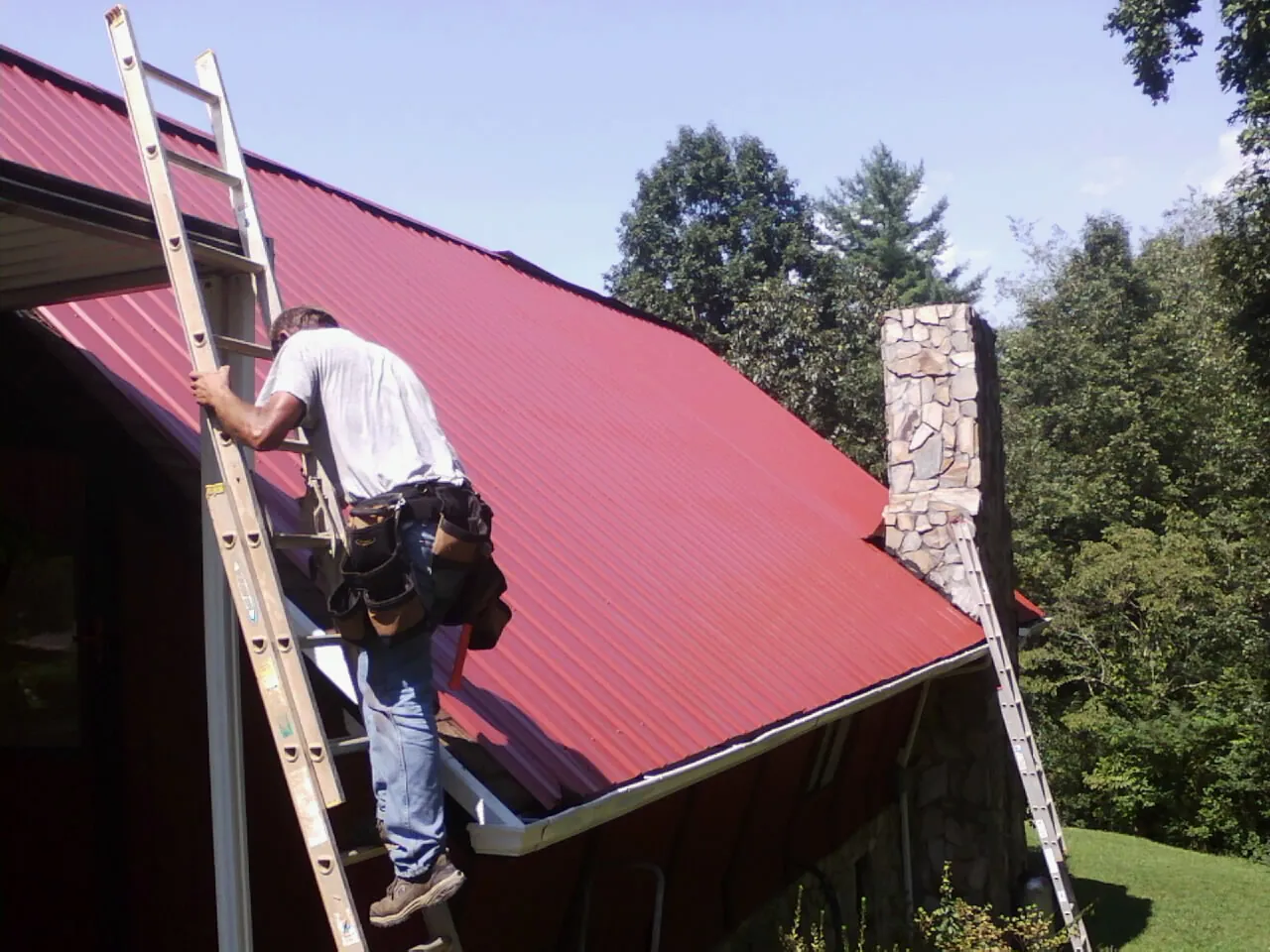 Roofer on a ladder beside a red metal roof and stone chimney — Soffit & Fascia Repair by Roofing Brothers in Crab Orchard